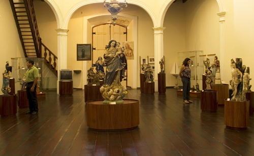 Fotografia da principal sala de exposição do Museu, com piso de madeira escura e colunas com arcos. Estão expostas esculturas sacras sobre bases de madeira, com destaque, ao centro, para uma grande imagem feminina com anjos aos seus pés. Há visitantes observando as obras e, no fundo da sala, uma escada de madeira à esquerda.
