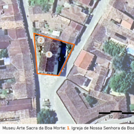 Imagem aérea do entorno do museu. No centro, uma área triangular está demarcada por uma linha laranja e contém dois elementos numerados: "1" Igreja de Nossa Senhora da Boa Morte e “2” anexo. A edificação marcada com o número 1 ocupa a maior parte da área demarcada. Há vegetação nas proximidades. A imagem inclui a legenda com a identificação das edificações, no canto inferior direito a escala gráfica e, na parte superior esquerda, uma seta indicando o ponto cardeal norte.
