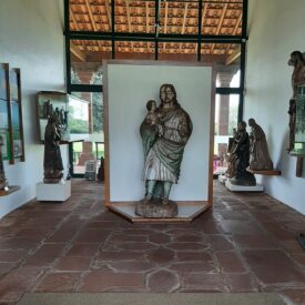 Fotografia de sala de exposição com esculturas, com destaque para uma escultura central de São José segurando uma criança. O ambiente possui paredes brancas e piso de pedra arenito vermelha. Ao fundo, é possível visualizar grandes esquadrias com vista para a área gramada.