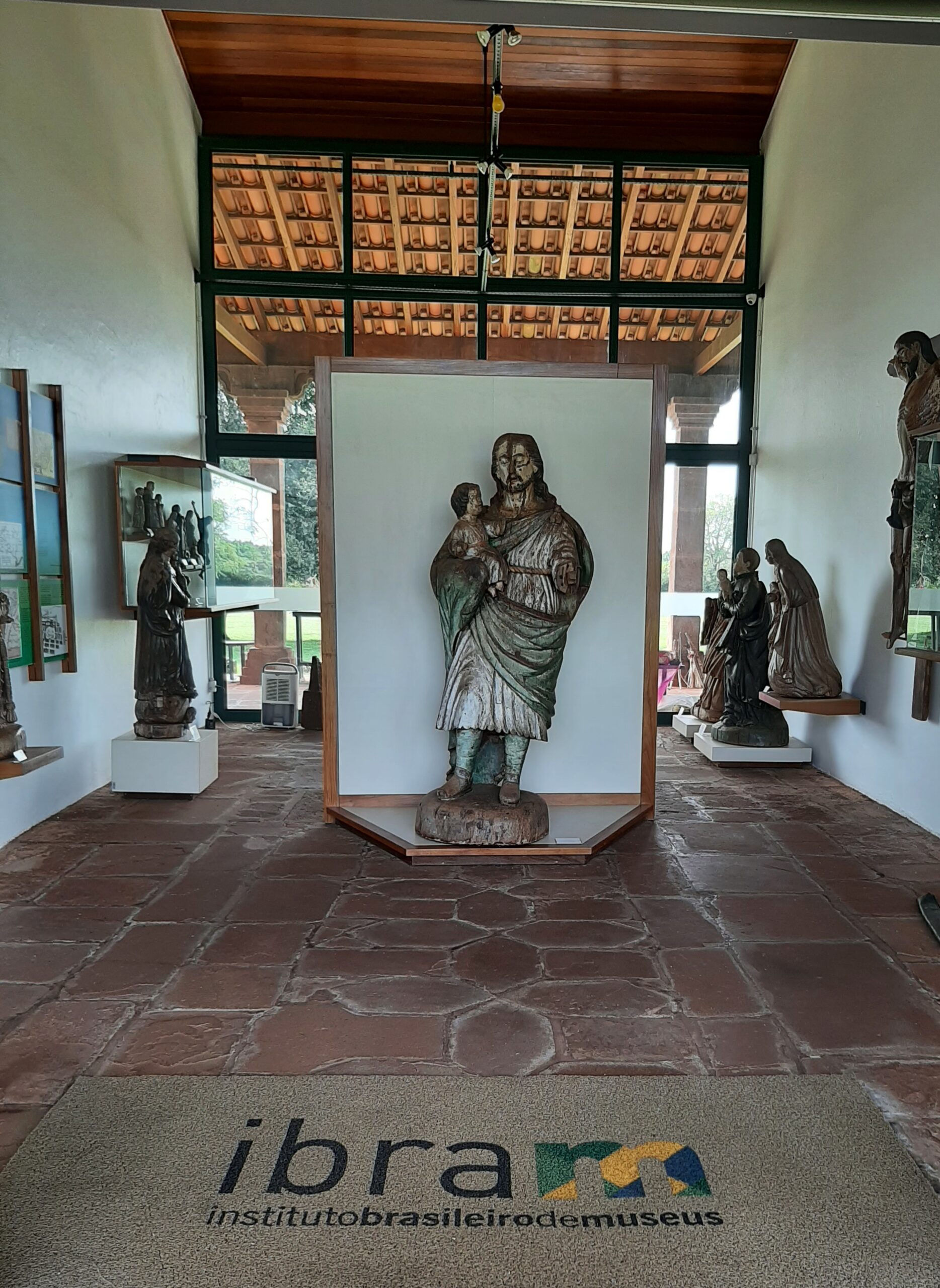Fotografia de sala de exposição com esculturas, com destaque para uma escultura central de São José segurando uma criança. O ambiente possui paredes brancas e piso de pedra arenito vermelha. Ao fundo, é possível visualizar grandes esquadrias com vista para a área gramada.