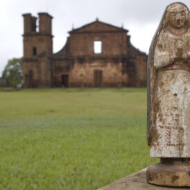 Fotografia da área gramada do Museu, apresentando uma estátua de figura religiosa em destaque, posicionada em primeiro plano. Ao fundo, de forma desfocada, é possível observar as ruínas da Igreja São Miguel Arcanjo, compondo um cenário que mistura elementos históricos e religiosos.