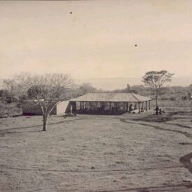 Fotografia histórica do Museu, em tons de sépia. Trata-se de construção térrea com telhado de quatro águas. A cobertura forma varandas cobertas, que são sustentadas por pilares. A edificação está localizada em um terreno amplo e gramado, com árvores ao fundo e paisagem rural ao redor. Algumas pessoas estão sentadas e em pé próximas à estrutura.