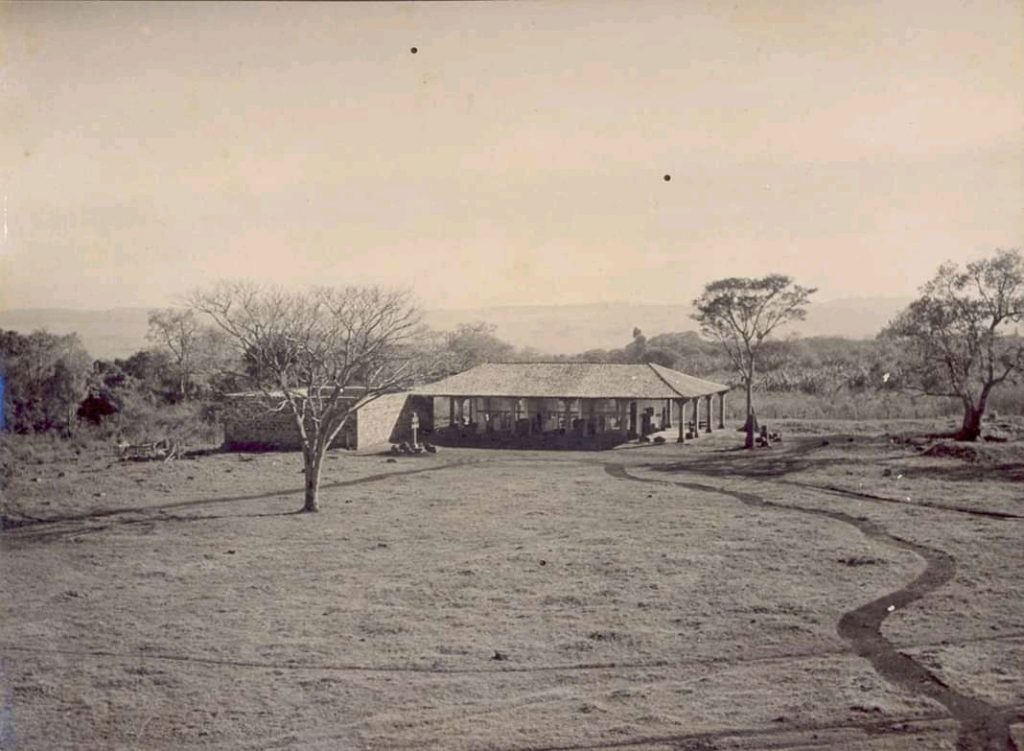 Fotografia histórica do Museu, em tons de sépia. Trata-se de construção térrea com telhado de quatro águas. A cobertura forma varandas cobertas, que são sustentadas por pilares. A edificação está localizada em um terreno amplo e gramado, com árvores ao fundo e paisagem rural ao redor. Algumas pessoas estão sentadas e em pé próximas à estrutura.