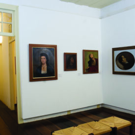 Fotografia de uma sala de exposição, onde quatro pinturas, variadas em tamanho e tipo de moldura, estão fixadas em uma parede branca. No centro da sala, há um banco de madeira com assento em fibras trançadas. O piso é de madeira escura. À esquerda, uma porta aberta dá acesso a outra sala expositiva, onde se nota outro quadro pendurado.