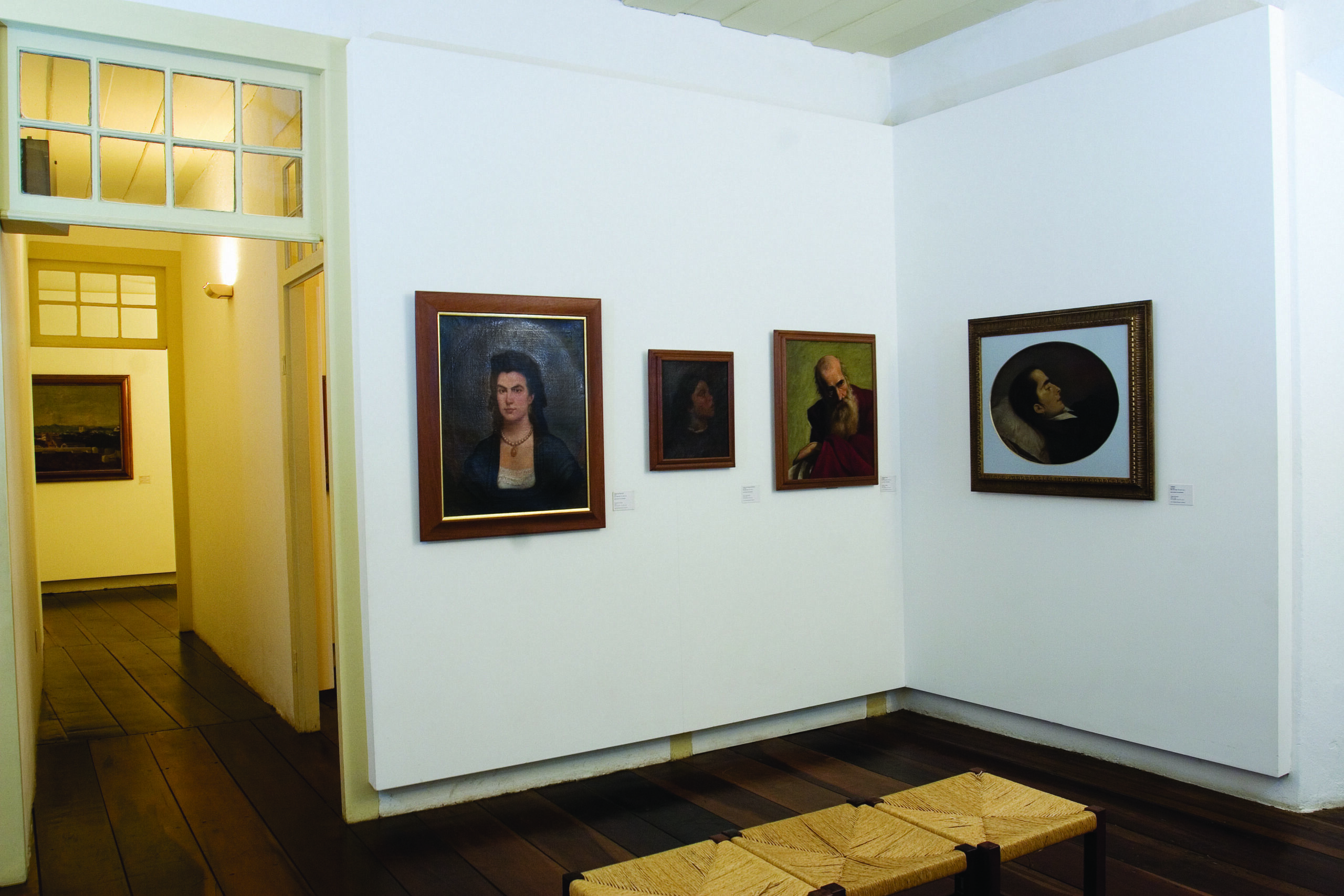 Fotografia de uma sala de exposição, onde quatro pinturas, variadas em tamanho e tipo de moldura, estão fixadas em uma parede branca. No centro da sala, há um banco de madeira com assento em fibras trançadas. O piso é de madeira escura. À esquerda, uma porta aberta dá acesso a outra sala expositiva, onde se nota outro quadro pendurado.