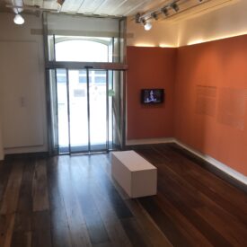 Fotografia de espaço expositivo com paredes pintadas na cor laranja e piso de madeira. À esquerda, observa-se uma obra de arte emoldurada e fixada na parede. No centro da sala, há um banco branco de formato retangular. Na parede do fundo, à direita de uma janela, encontra-se uma televisão que exibe um vídeo, acompanhada por um texto explicativo fixado na parede ao lado.