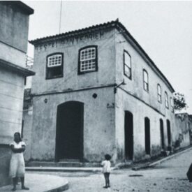 Fotografia histórica em preto e branco do museu, edifício de dois andares localizado na esquina de duas vias, com janelas e portas retangulares e verga em arco abatido. Em frente ao edifício, uma criança pequena está de pé na calçada, olhando para o prédio.