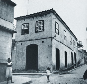 Fotografia histórica em preto e branco do museu, edifício de dois andares localizado na esquina de duas vias, com janelas e portas retangulares e verga em arco abatido. Em frente ao edifício, uma criança pequena está de pé na calçada, olhando para o prédio.