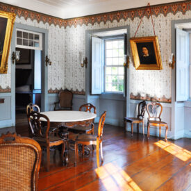 Fotografia de espaço expositivo, com paredes decoradas por papel de parede de influência estética clássica e detalhes em dourado, e o piso do ambiente em madeira. No centro, há uma mesa redonda de madeira escura cercada por cadeiras. Ao fundo, estão dois retratos emoldurados, um fixado na parede à esquerda e outro à direita, ambos com molduras douradas. No lado direito, há duas janelas abertas.