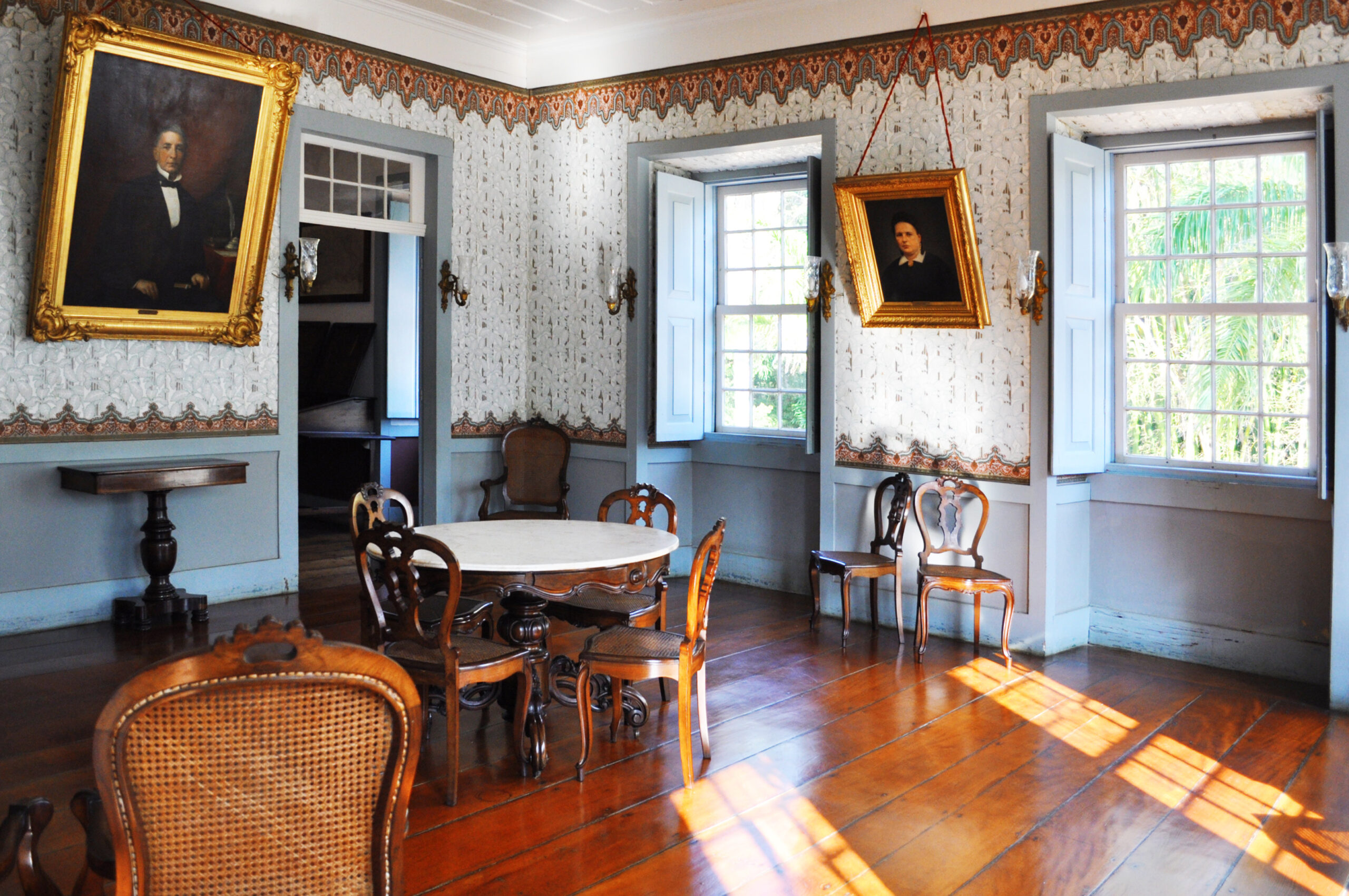 Fotografia de espaço expositivo, com paredes decoradas por papel de parede de influência estética clássica e detalhes em dourado, e o piso do ambiente em madeira. No centro, há uma mesa redonda de madeira escura cercada por cadeiras. Ao fundo, estão dois retratos emoldurados, um fixado na parede à esquerda e outro à direita, ambos com molduras douradas. No lado direito, há duas janelas abertas.