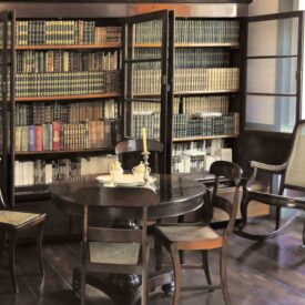 Fotografia do espaço expositivo da biblioteca do museu. O espaço possui estantes de livros ao fundo e, em primeiro plano, ao centro, uma mesa redonda de madeira com quatro cadeiras e objetos decorativos. À direita, destaca-se uma cadeira de balanço junto à janela. O piso é de madeira e a luz natural ilumina o espaço através da janela lateral.