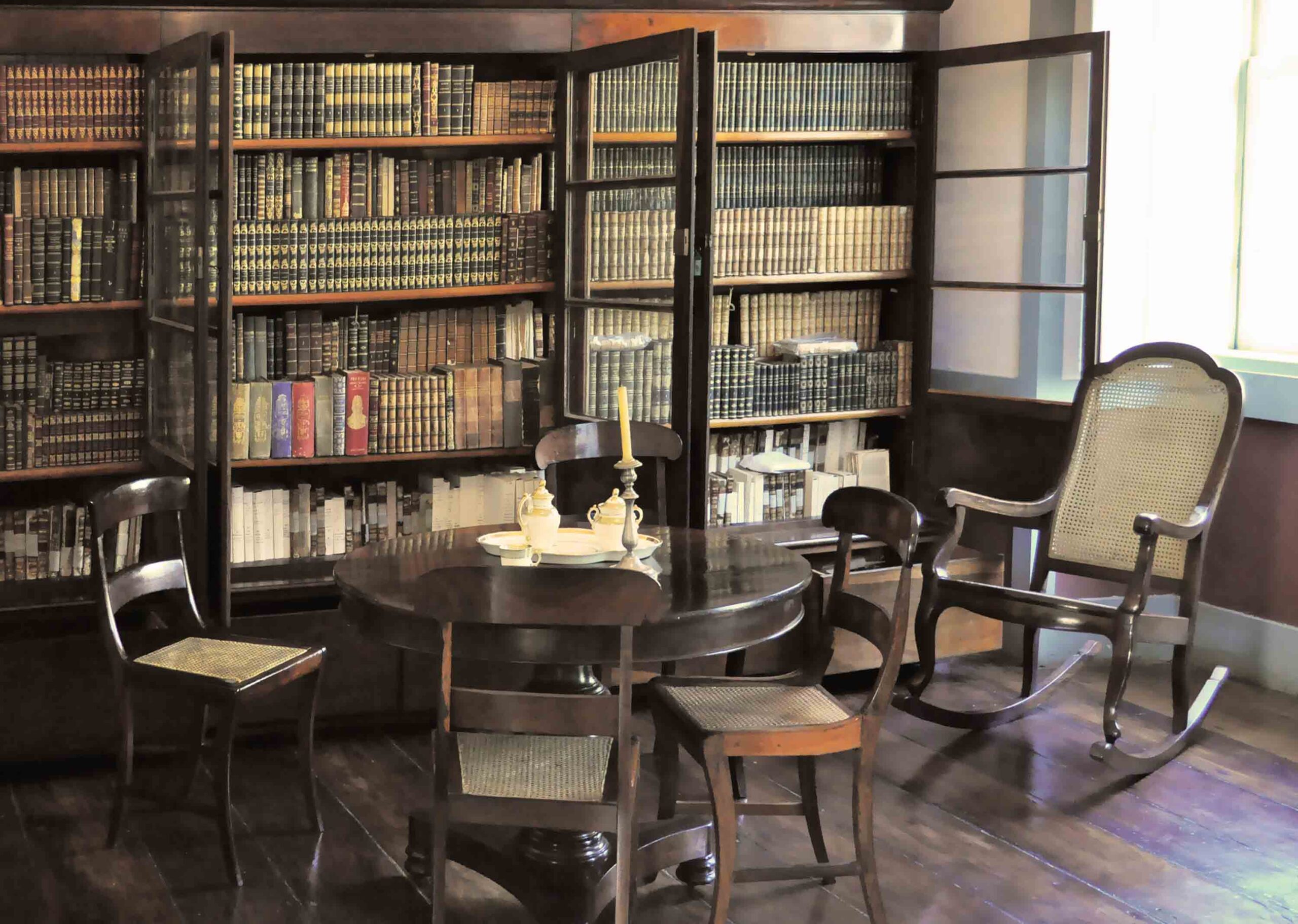 Fotografia do espaço expositivo da biblioteca do museu. O espaço possui estantes de livros ao fundo e, em primeiro plano, ao centro, uma mesa redonda de madeira com quatro cadeiras e objetos decorativos. À direita, destaca-se uma cadeira de balanço junto à janela. O piso é de madeira e a luz natural ilumina o espaço através da janela lateral.