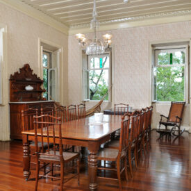 Imagem da sala de jantar, área expositiva do Museu. No centro da sala, há uma grande mesa de jantar de madeira acompanhada de 12 cadeiras de madeira. A sala possui paredes claras com um sutil padrão floral, teto branco e piso de madeira. Ao fundo, há duas janelas em uma das paredes, ladeadas no canto direito por estante de louças. Na parede esquerda, há outro móvel louceiro posicionado entre duas janelas. Por meio das janelas, observa-se a vegetação ao redor do edifício.