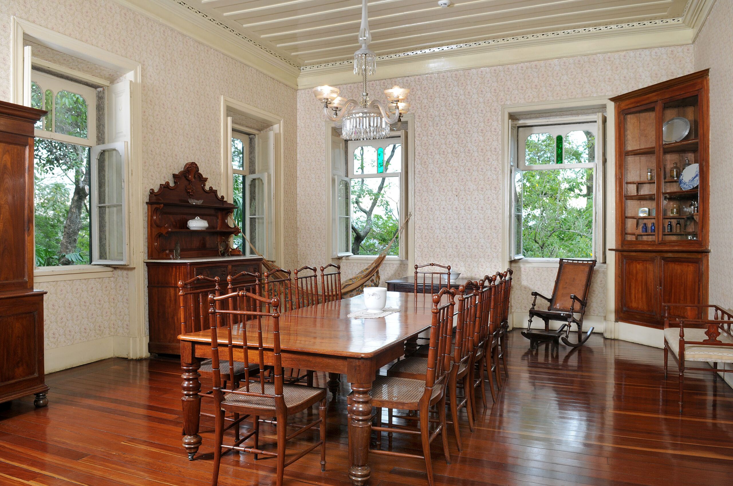 Imagem da sala de jantar, área expositiva do Museu. No centro da sala, há uma grande mesa de jantar de madeira acompanhada de 12 cadeiras de madeira. A sala possui paredes claras com um sutil padrão floral, teto branco e piso de madeira. Ao fundo, há duas janelas em uma das paredes, ladeadas no canto direito por estante de louças. Na parede esquerda, há outro móvel louceiro posicionado entre duas janelas. Por meio das janelas, observa-se a vegetação ao redor do edifício.