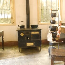 Imagem da cozinha, sala expositiva do museu. O espaço possui paredes brancas e piso decorado com ladrilhos hidráulicos. O fogão a lenha é o ponto focal central, com uma mesa no canto esquerdo, um banquinho em frente ao fogão e outros utensílios de cozinha pendurados em um suporte metálico. A iluminação é de luz natural proveniente de duas janelas envidraçadas que ladeiam o fogão.