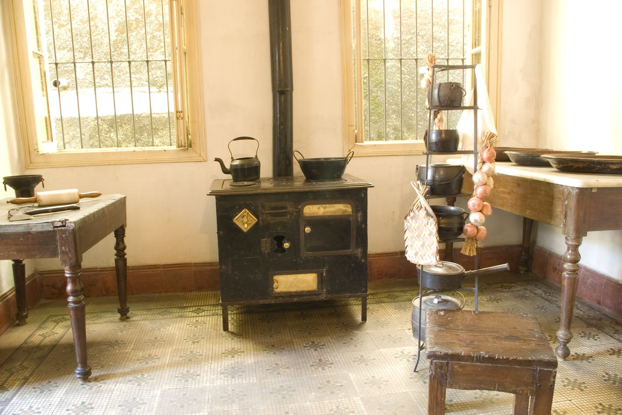 Imagem da cozinha, sala expositiva do museu. O espaço possui paredes brancas e piso decorado com ladrilhos hidráulicos. O fogão a lenha é o ponto focal central, com uma mesa no canto esquerdo, um banquinho em frente ao fogão e outros utensílios de cozinha pendurados em um suporte metálico. A iluminação é de luz natural proveniente de duas janelas envidraçadas que ladeiam o fogão.
