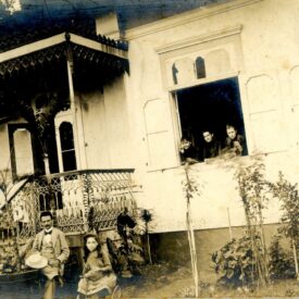 Fotografia antiga em preto e branco, com uma família reunida em frente à casa principal. Na imagem, é possível ver três mulheres nas janelas, enquanto dois homens e uma criança estão sentados em frente à varanda da casa. A imagem mostra a casa, atual Museu Casa de Benjamin Constant.