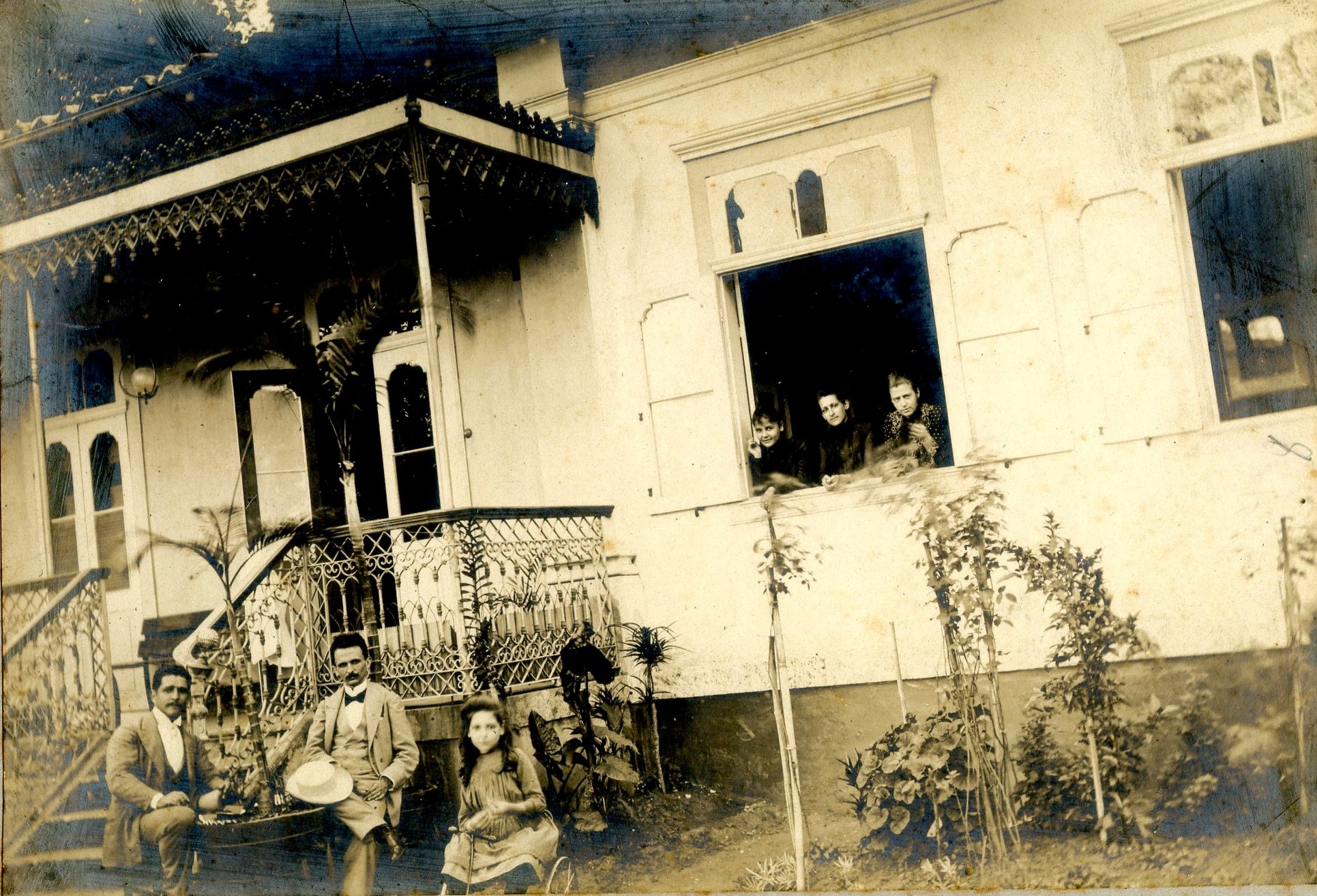 Fotografia antiga em preto e branco, com uma família reunida em frente à casa principal. Na imagem, é possível ver três mulheres nas janelas, enquanto dois homens e uma criança estão sentados em frente à varanda da casa. A imagem mostra a casa, atual Museu Casa de Benjamin Constant.