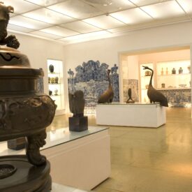 Fotografia do espaço de exposição. No primeiro plano à esquerda, há um objeto de bronze com relevos. No fundo, há esculturas de vários tamanhos, incluindo figuras de pássaros em pedestais brancos. Os painéis de azulejos azuis com cenas figurativas revestem as paredes ao fundo. Na parede à direita, existem nichos embutidos onde estão expostas peças menores.