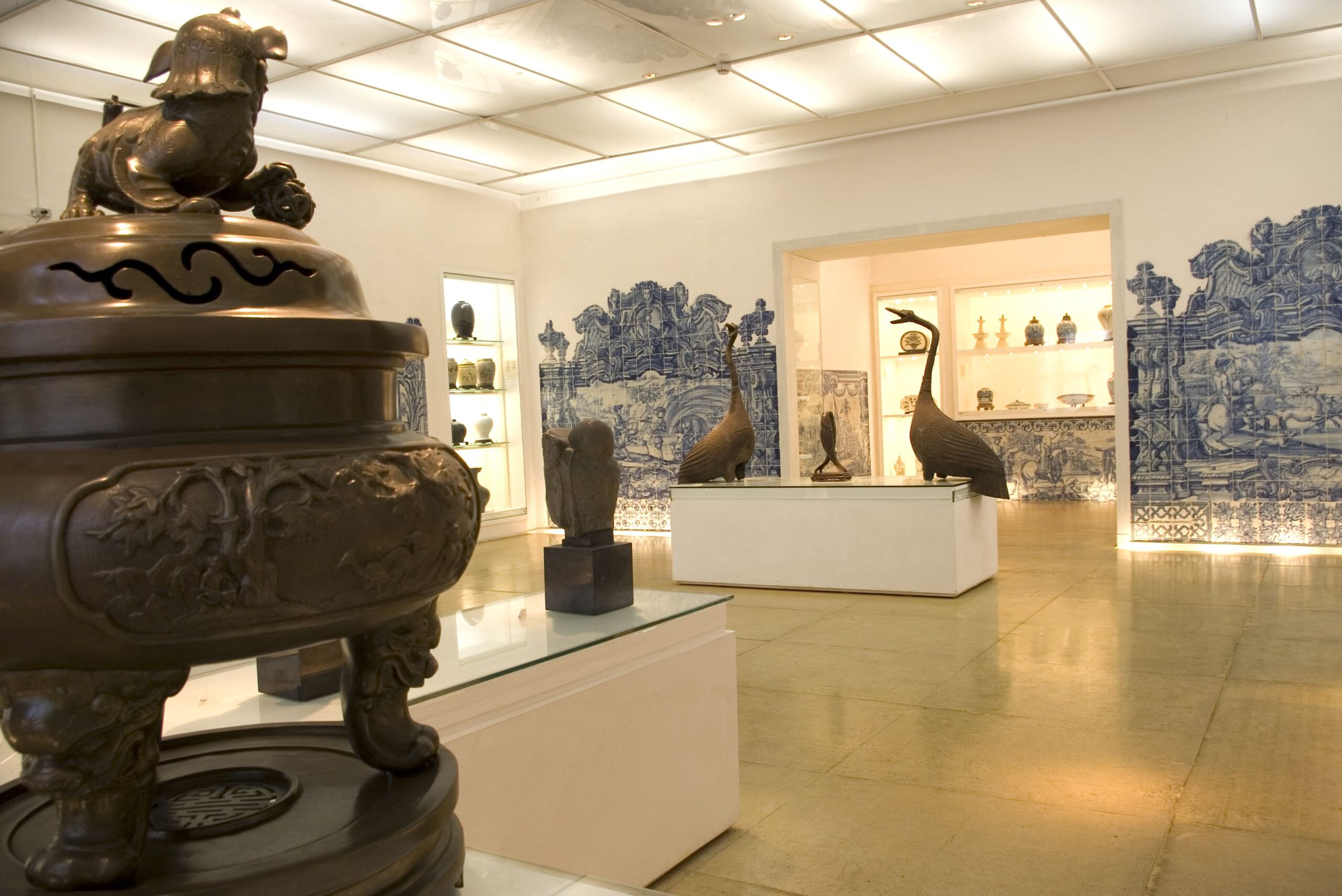 Fotografia do espaço de exposição. No primeiro plano à esquerda, há um objeto de bronze com relevos. No fundo, há esculturas de vários tamanhos, incluindo figuras de pássaros em pedestais brancos. Os painéis de azulejos azuis com cenas figurativas revestem as paredes ao fundo. Na parede à direita, existem nichos embutidos onde estão expostas peças menores.