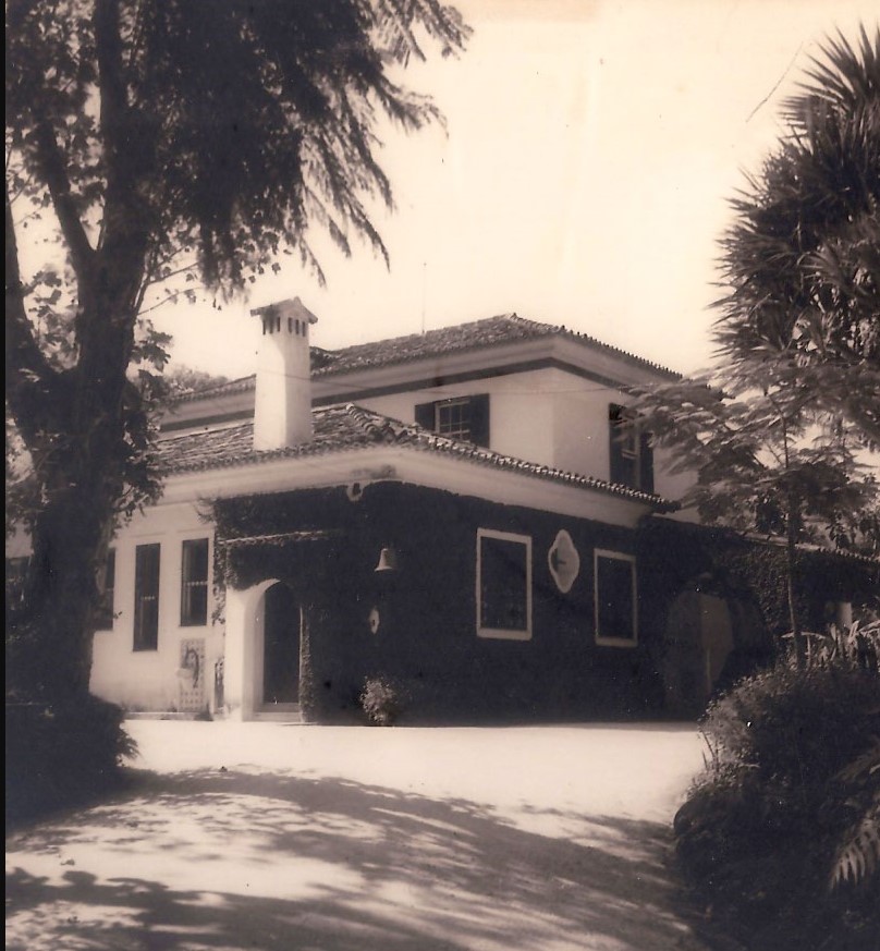 Fotografia antiga do museu, em preto e branco. O edifício tem dois andares, sendo que o andar superior é menor que o térreo. A cobertura é em telhas cerâmicas com quatro águas. No andar térreo, à esquerda, há uma porta em arco, e ao longo da fachada estão dispostas diversas janelas retangulares. Uma parte da parede térrea está revestida por trepadeiras. Árvores e vegetação cercam o edifício, criando sombras no chão à sua frente.