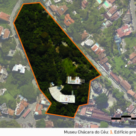 Vista aérea de uma área urbana, destacando um grande terreno contornado por uma linha laranja. Dentro desse perímetro, há duas construções identificadas com os números "1" e "2". A construção "1", situada na parte norte do terreno, é o edifício principal do museu, enquanto a construção "2", localizada mais ao sul, é o anexo. A imagem inclui a legenda com a identificação das edificações, no canto inferior direito a escala gráfica e, na parte superior esquerda, uma seta indicando o ponto cardeal norte.
