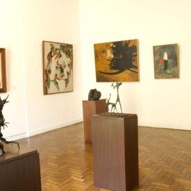 Imagem do espaço expositivo interno, ambiente com piso de madeira e paredes brancas. No primeiro plano, duas esculturas sobre pedestais de madeira se destacam: à esquerda, uma obra que sugere uma figura humana em movimento e, à direita, uma escultura de natureza mais abstrata. Ao fundo, há quatro pinturas de estilos e tamanhos variados expostas nas paredes.