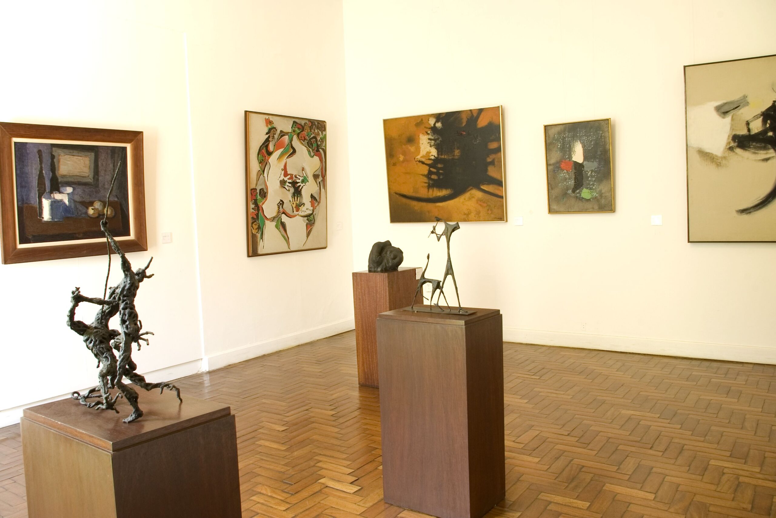 Imagem do espaço expositivo interno, ambiente com piso de madeira e paredes brancas. No primeiro plano, duas esculturas sobre pedestais de madeira se destacam: à esquerda, uma obra que sugere uma figura humana em movimento e, à direita, uma escultura de natureza mais abstrata. Ao fundo, há quatro pinturas de estilos e tamanhos variados expostas nas paredes.