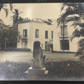 Registro fotográfico histórico do museu em preto e branco. A edificação de dois andares conta com várias janelas e portas de vidro, algumas delas com varandas. Em destaque, no jardim, há uma estátua de um leão sentado sobre um pedestal e ao redor, diferentes vegetações.