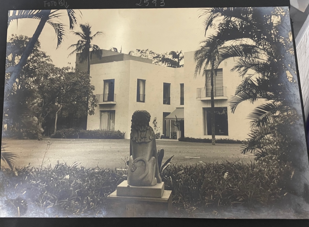 Registro fotográfico histórico do museu em preto e branco. A edificação de dois andares conta com várias janelas e portas de vidro, algumas delas com varandas. Em destaque, no jardim, há uma estátua de um leão sentado sobre um pedestal e ao redor, diferentes vegetações.