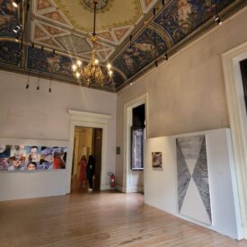 Fotografia da sala de exposições do Museu, com várias peças de arte em exibição, incluindo fotografias e pinturas abstratas. A sala possui paredes brancas com painéis de exposições ao longo das paredes, piso de madeira clara e teto decorado. As janelas estão abertas e são voltadas para o jardim. No teto, um grande lustre colocado no centro ilumina o local.