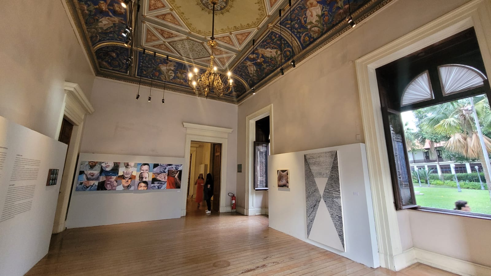 Fotografia da sala de exposições do Museu, com várias peças de arte em exibição, incluindo fotografias e pinturas abstratas. A sala possui paredes brancas com painéis de exposições ao longo das paredes, piso de madeira clara e teto decorado. As janelas estão abertas e são voltadas para o jardim. No teto, um grande lustre colocado no centro ilumina o local.