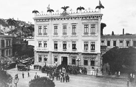 Fotografia histórica em preto e branco da fachada do Museu da República e seu entorno, com destaque para a praça localizada em frente ao edifício, onde um grande grupo de pessoas está presente. A rua está repleta de pessoas e veículos, indicando intensa atividade e importância do local.