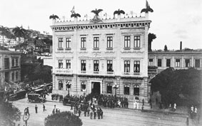 Fotografia histórica em preto e branco da fachada do Museu da República e seu entorno, com destaque para a praça localizada em frente ao edifício, onde um grande grupo de pessoas está presente. A rua está repleta de pessoas e veículos, indicando intensa atividade e importância do local.