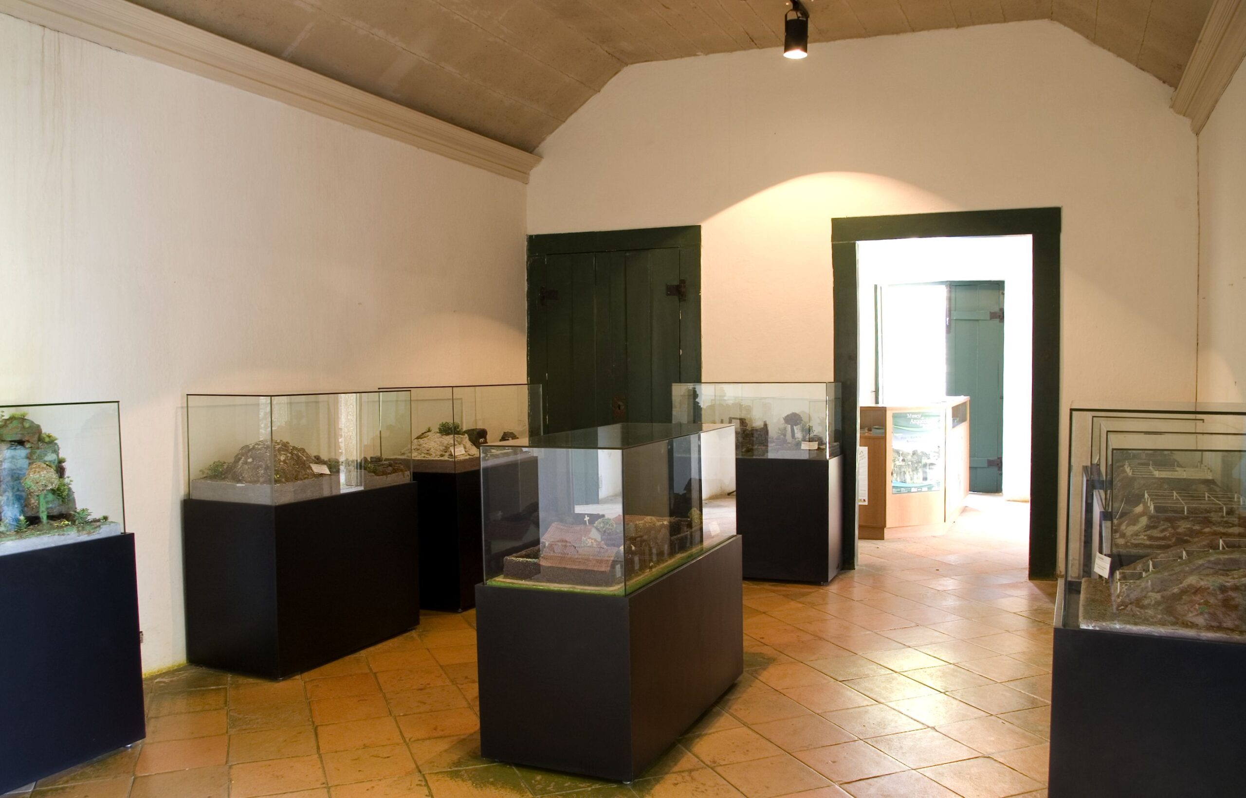 Sala expositiva, retangular, com paredes, teto e piso claros. O forro do teto é arqueado e a sala possui diversas vitrines com objetos expostos. As vitrines possuem fechamento em vidro, apoiadas sobre suportes pretos. Em segundo plano, nota-se um vão que leva a outro ambiente que dá continuidade à exposição.