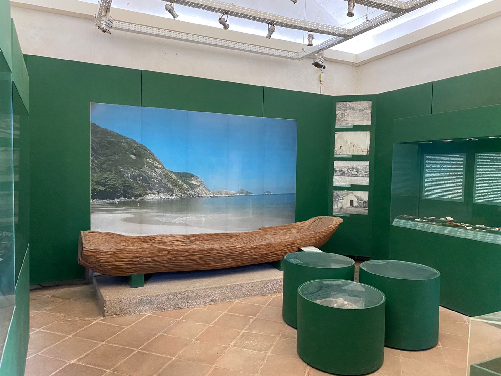 Fotografia de espaço expositivo, com paredes pintadas de verde, teto claro e piso em cerâmica clara. No centro da sala existem três vitrines redondas, baixas, contendo objetos em destaque. Na parede do fundo, um quadro com paisagem de uma praia e uma canoa de madeira exposta sobre um suporte. À direita da sala, há nichos expositores com artefatos e textos explicativos.