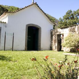 Fotografia da fachada da capela em perspectiva. Observa-se a cobertura em telhas cerâmicas e telhado de duas águas, com uma cruz no topo, simbolizando o uso religioso. A entrada é emoldurada por um arco abatido de pedras centralizado na fachada. À direita, um muro de pedras com uma abertura leva a outra parte do espaço externo. Em frente à capela, há um extenso jardim com flores.
