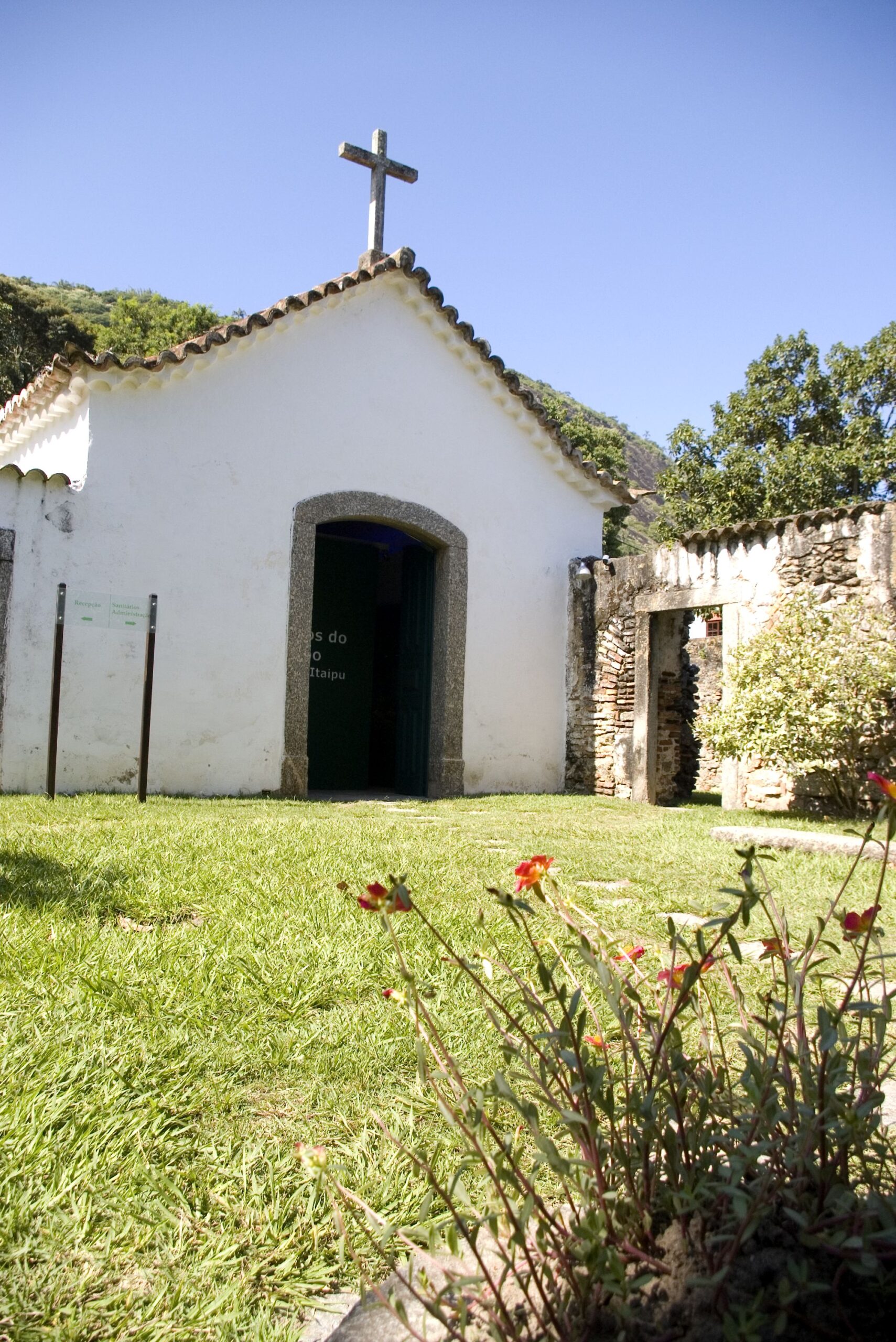 Fotografia da fachada da capela em perspectiva. Observa-se a cobertura em telhas cerâmicas e telhado de duas águas, com uma cruz no topo, simbolizando o uso religioso. A entrada é emoldurada por um arco abatido de pedras centralizado na fachada. À direita, um muro de pedras com uma abertura leva a outra parte do espaço externo. Em frente à capela, há um extenso jardim com flores.