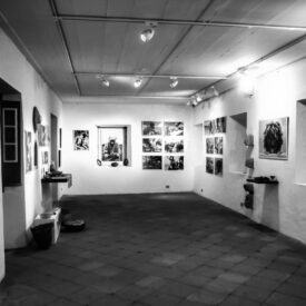 Imagem em preto e branco do espaço de exposição do museu. O local conta com paredes e teto claros, iluminação direcionada por trilhos e vários quadros com imagens que representam a exposição. O piso é de cerâmica.