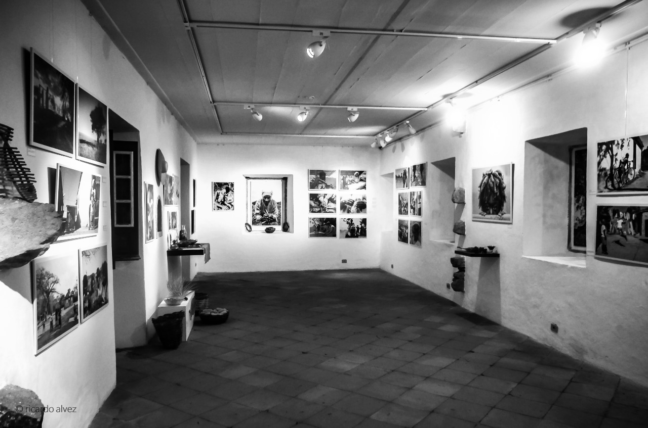 Imagem em preto e branco do espaço de exposição do museu. O local conta com paredes e teto claros, iluminação direcionada por trilhos e vários quadros com imagens que representam a exposição. O piso é de cerâmica.