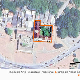 Vista de satélite da área do museu e seu entorno, mostrando a malha urbana da área que circunda o museu. No centro da imagem, um retângulo laranja destaca a igreja de Nossa Senhora dos Anjos, número 1, e o salão do museu, número 2, situados próximos a uma área verde. Abaixo da imagem, há uma legenda identificando os elementos como parte do Museu de Arte Religiosa e Tradicional. A imagem inclui a legenda com a identificação das edificações, no canto inferior direito a escala gráfica e, na parte superior esquerda, uma seta indicando o ponto cardeal norte.