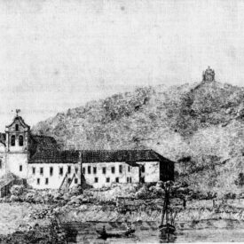 Fotografia histórica em preto e branco do museu. O conjunto de edifícios é composto por volumes de diferentes alturas e formatos. À esquerda da imagem, observa-se a igreja com a torre lateral e, à direita, uma edificação retangular de dois pavimentos com diversas janelas distribuídas ao longo da fachada. Ao fundo, observa-se uma colina coberta por vegetação.