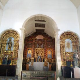 Fotografia interna do altar da igreja, composta por três nichos ornamentados. O nicho central é o maior e mais elaborado, com detalhes dourados e diversas imagens religiosas. Os nichos laterais, menores, também abrigam imagens religiosas cercadas por ornamentos dourados.