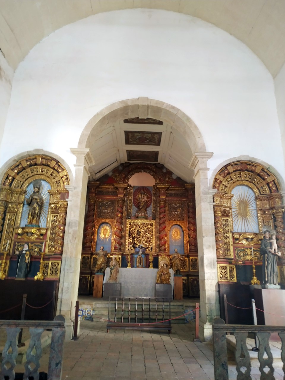 Fotografia interna do altar da igreja, composta por três nichos ornamentados. O nicho central é o maior e mais elaborado, com detalhes dourados e diversas imagens religiosas. Os nichos laterais, menores, também abrigam imagens religiosas cercadas por ornamentos dourados.
