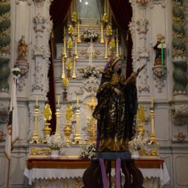 Fotografia do altar da igreja com a imagem de Nossa Senhora das Dores ao centro, vestida com manto roxo escuro e detalhes dourados. O fundo do altar é decorado com esculturas e cortinas vermelhas que emolduram a parte superior.