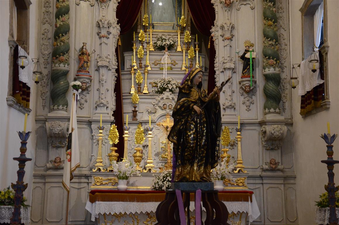 Fotografia do altar da igreja com a imagem de Nossa Senhora das Dores ao centro, vestida com manto roxo escuro e detalhes dourados. O fundo do altar é decorado com esculturas e cortinas vermelhas que emolduram a parte superior.