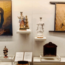 Fotografia da vitrine de exposição com imagens sacras e objetos religiosos. Ao fundo, duas pinturas de santas emolduradas, uma à esquerda e outra à direita. Ao centro, há cinco esculturas religiosas de diferentes tamanhos, representando figuras católicas, dispostas em suportes de vidro e prateleiras brancas. A iluminação destaca cada item individualmente.
