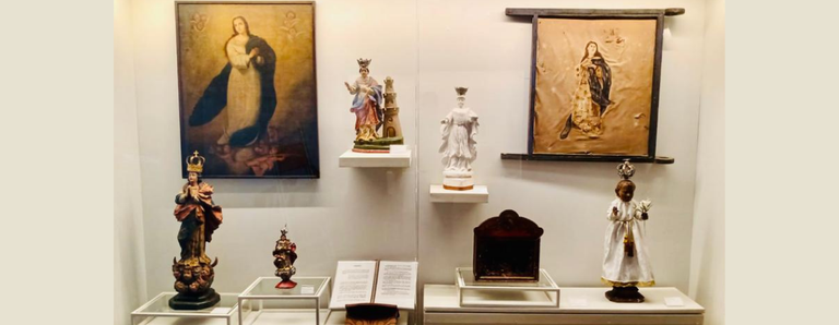 Fotografia da vitrine de exposição com imagens sacras e objetos religiosos. Ao fundo, duas pinturas de santas emolduradas, uma à esquerda e outra à direita. Ao centro, há cinco esculturas religiosas de diferentes tamanhos, representando figuras católicas, dispostas em suportes de vidro e prateleiras brancas. A iluminação destaca cada item individualmente.