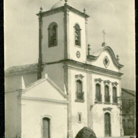 Fotografia histórica em preto e branco da fachada da Igreja, localizada de frente para uma praça. A imagem evidencia a arquitetura colonial da igreja e sua capela lateral, em um ambiente vazio, sem presença de pessoas ou veículos.