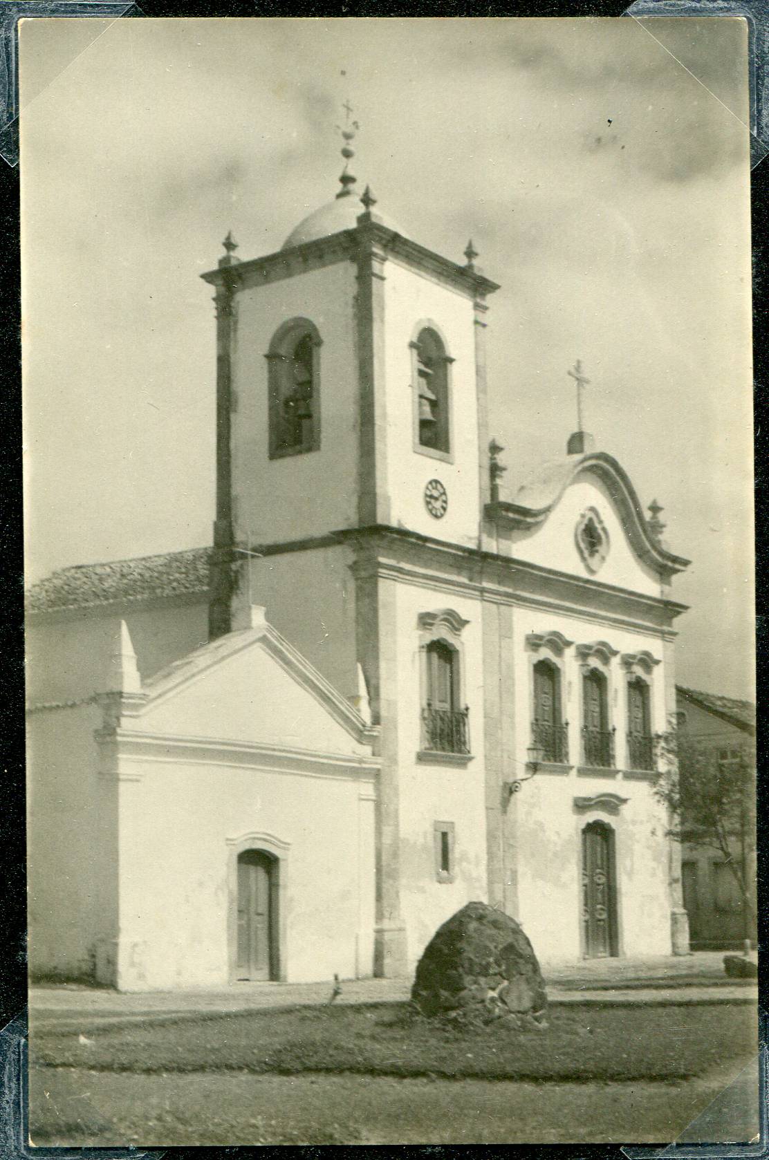 Fotografia histórica em preto e branco da fachada da Igreja, localizada de frente para uma praça. A imagem evidencia a arquitetura colonial da igreja e sua capela lateral, em um ambiente vazio, sem presença de pessoas ou veículos.